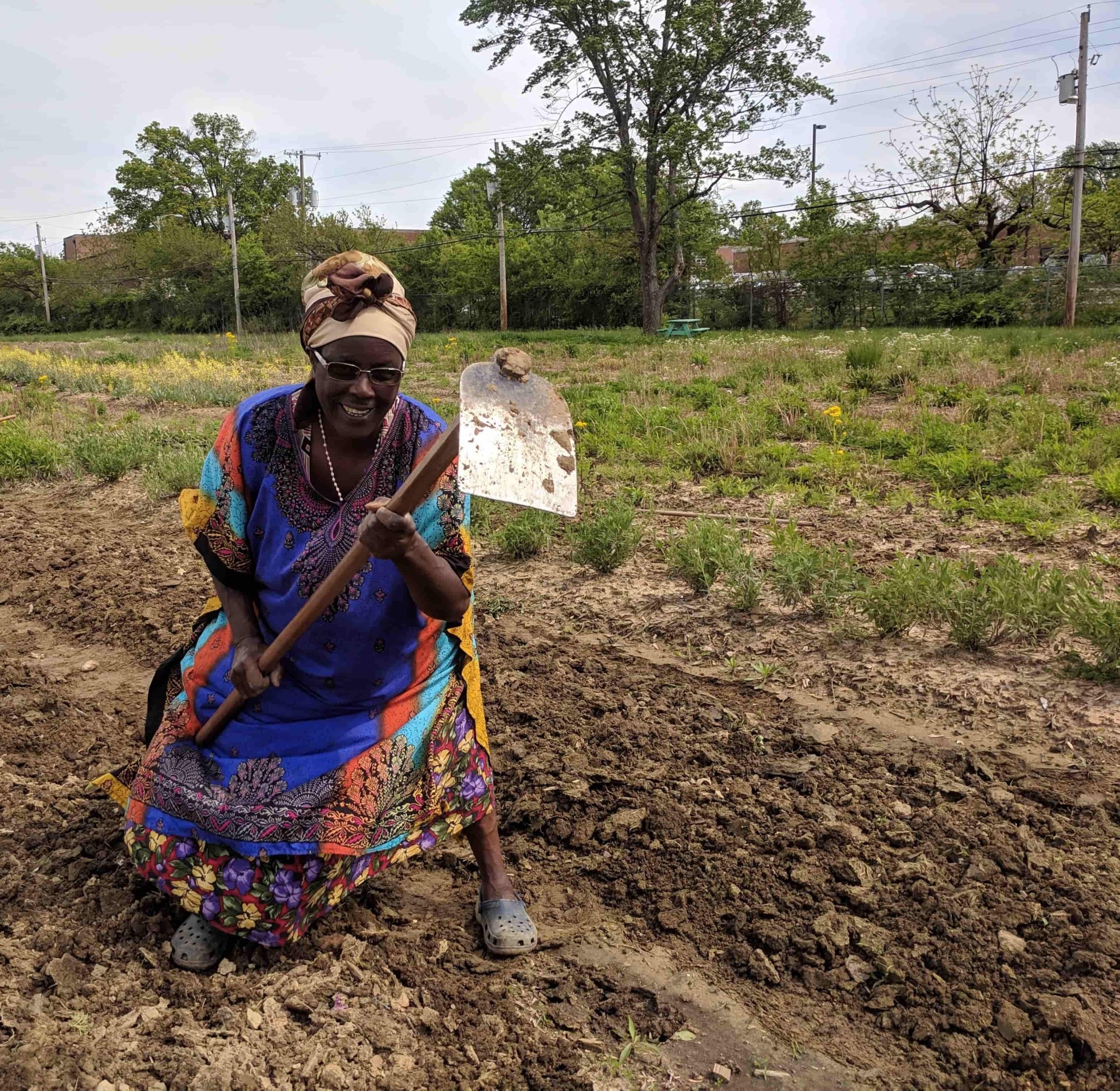 An elder woman in a bright dress kneeling in the soil with a hoe, smiling.